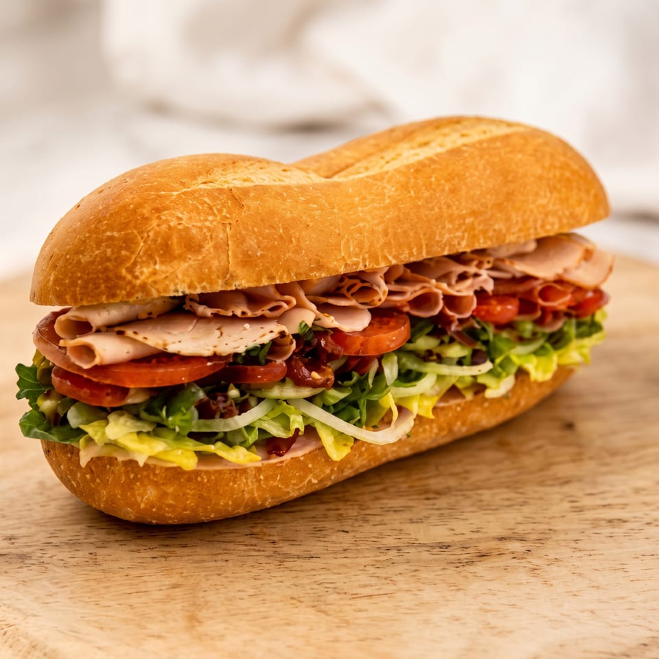 A large deli sandwich with turkey, tomatoes, lettuce, and onions on a wooden board.